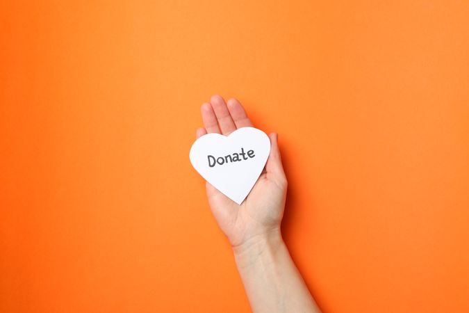 Female hand holds heart with inscription Donate on orange background, top view