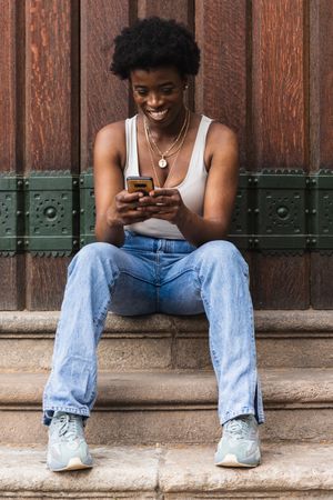 Smiling Black woman using smartphone sitting near building