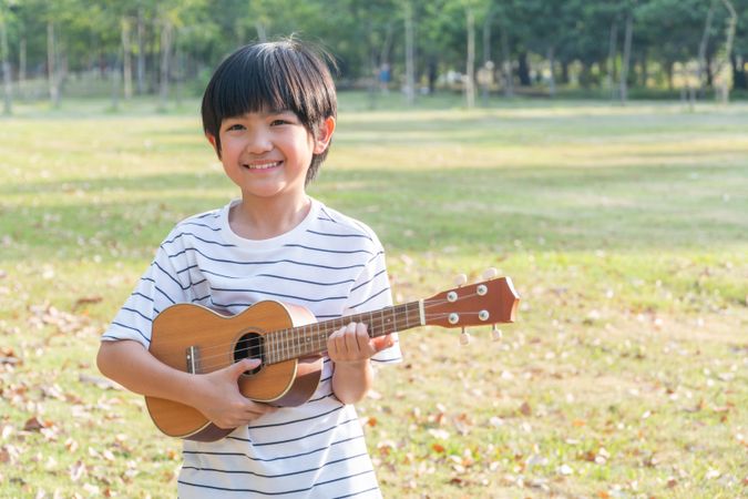 Smiling young boy playing ukulele in public park