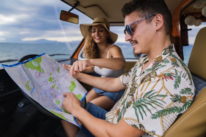 Touristic man exploring map with help of long haired woman in hat in front seat of car