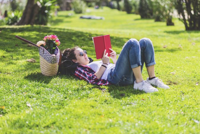 Side view of a hipster happy woman lying on grass in sunny day at park while reading a red book