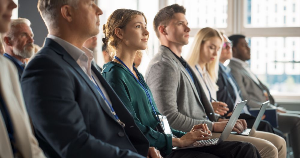 Young Woman Sitting in Crowded Audience at a Business Conference. Female Delegate Smiling, Using Laptop Computer. Manager Watching Motivational Presentation About Investing In Innovative Startups.