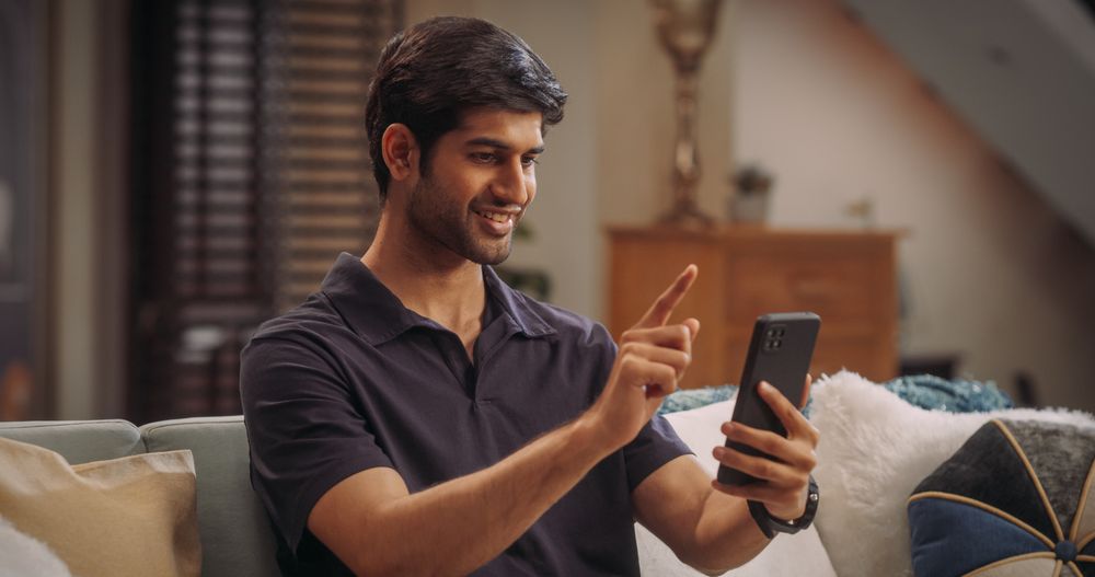 Cheerful Young Hindu Man Comfortably Using His Smartphone At Home, Handsome, Smiling and Positive