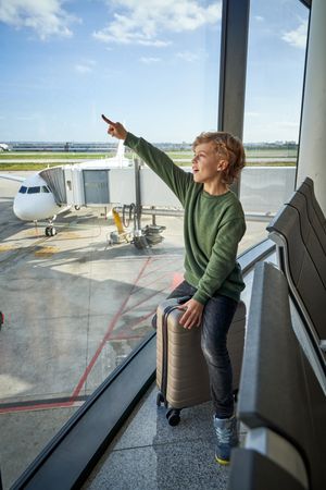 Happy kid pointing away sitting near window in airport terminal