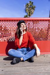 Happy woman in red sitting cross legged talking on mobile phone outdoors on a bright day