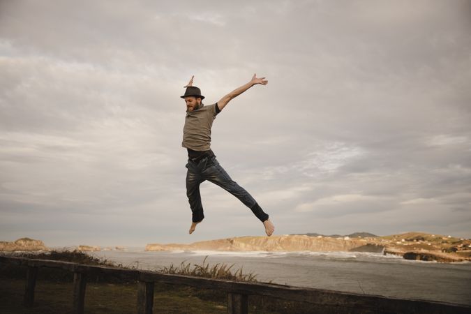 Young man in hats having fun on seat on coast near waving sea and cloudy sky