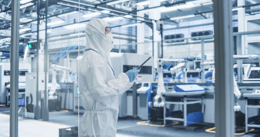 Female Technician in Protective Coveralls, Mask and Eyewear Using Tablet Computer at a Manufacturing Complex. Specialist Monitoring Conditions at a Modern Electronics Factory with Automated Robots