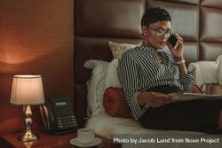 Businesswoman on hotel room bed reading newspaper and talking on cell phone 0Kyz14
