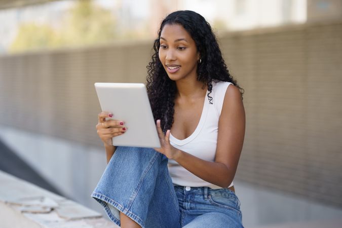A young woman is actively engaged with her digital tablet while sitting outdoors in the sunlight