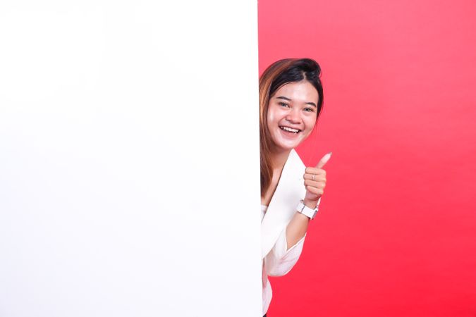 A woman with medium brown hair smiles, gives a thumbs up, and peeks from behind a large board, isolated on a red background.
