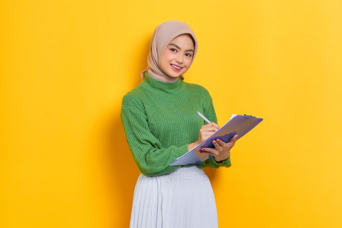 Smiling woman in headscarf writing on clipboard in studio shoot