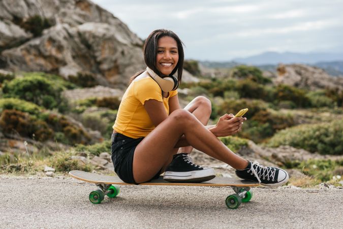 Hispanic cheerful woman using mobile phone while sitting on longboard