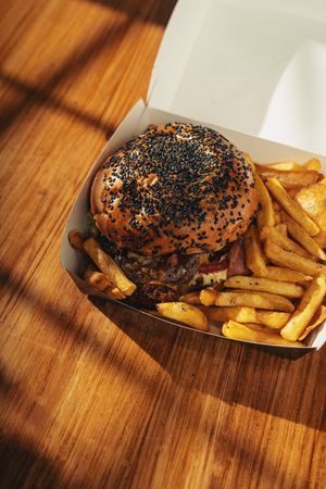 Gourmet burger with sesame bun and fries in takeout box on wooden table