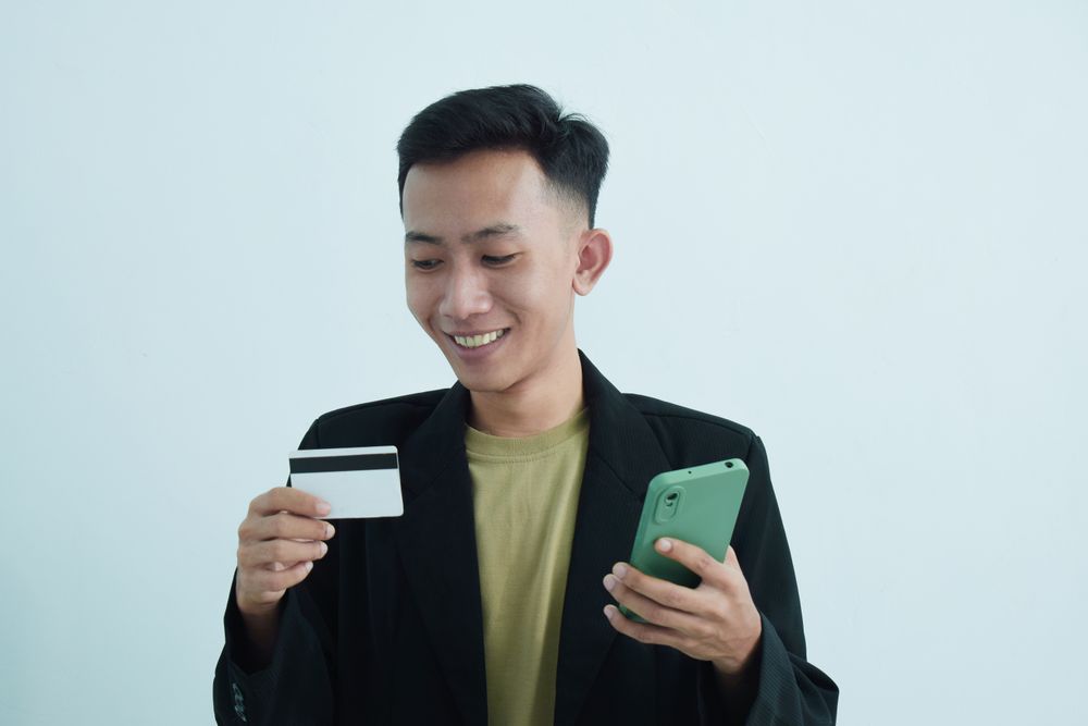 Smiling Asian businessman in suit holding blank credit card and smartphone