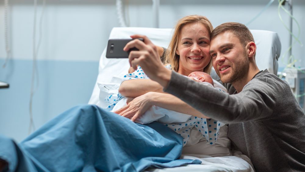 Father Takes Selfie of Him with His Wife Holding Newborn Baby while Lying on the Hospital Bed