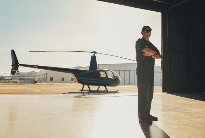 Man standing tall with arms crossed in helicopter hangar on sunny day