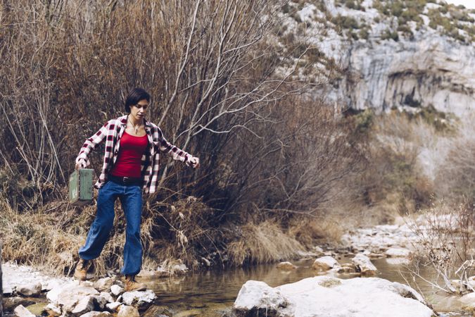 Woman with case jumping on rocks above stream