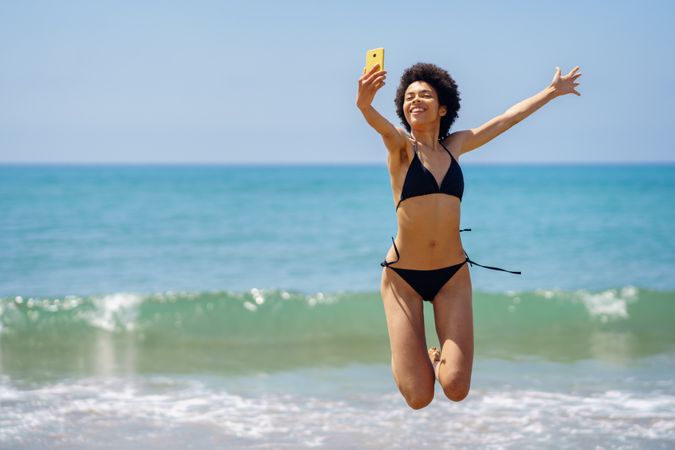 Woman in bikini happily jumping in front of the ocean and taking a selfie