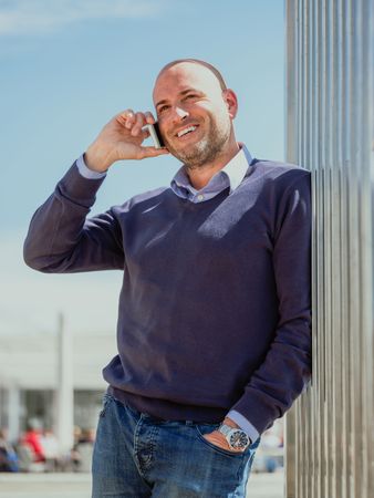 vertical photo of a businessman leaning on a wall of a building and talking to the mobile smiling