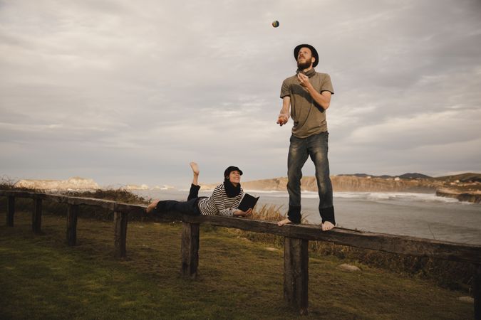 Man juggling balls on bench near woman with drum on shore