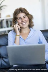 Smiling woman sitting on sofa at home using a laptop and chatting on phone 4AzJeq