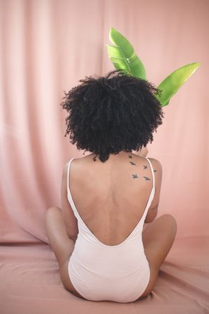 Back view of woman with afro hair wearing one piece bathing suit sitting on floor holding tree leaf in studio