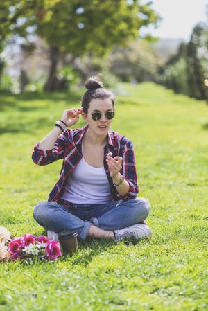 Front view of a young hipster woman sitting on grass in a park while holding a flower in a sunny day