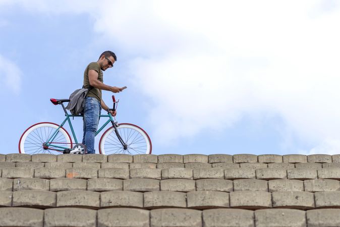 Male chatting on phone and standing with bike atop steps with beautiful blue sky and clouds