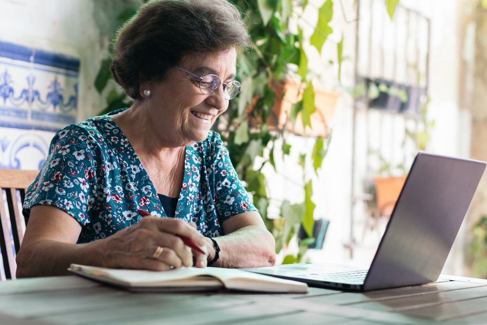 Older woman with glasses using a laptop and a notebook at home