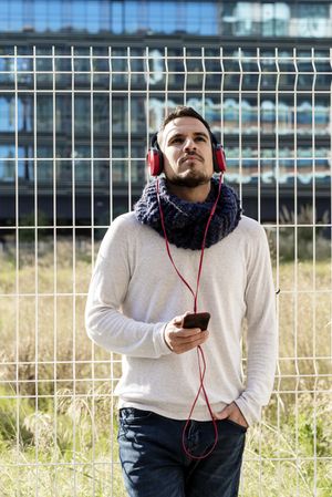 Young man walking in scarf leaning back on metallic fence and looking up from smartphone