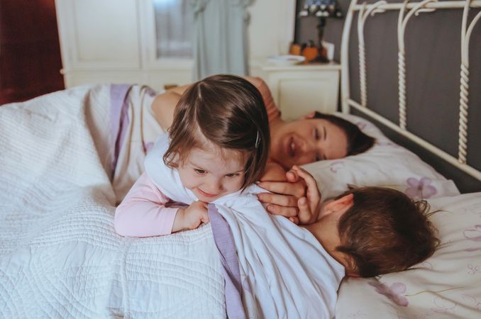 Portrait of adorable little girl playing over brother lying in the bed with mother