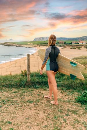 Female walking with surfboard atop of cliff with beautiful coastal view, vertical