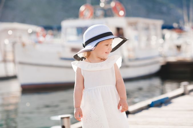Young girl in light dress with hat standing beside docked boats