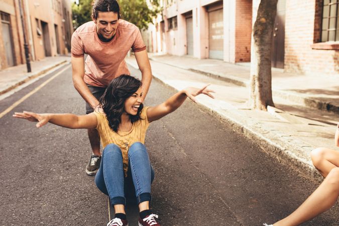 Excited young woman being pushing on skateboard by her boyfriend outdoors on street