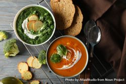 Two bowls of orange and green soup on wooden table with crackers, vegetables and spoons 0vJNG0