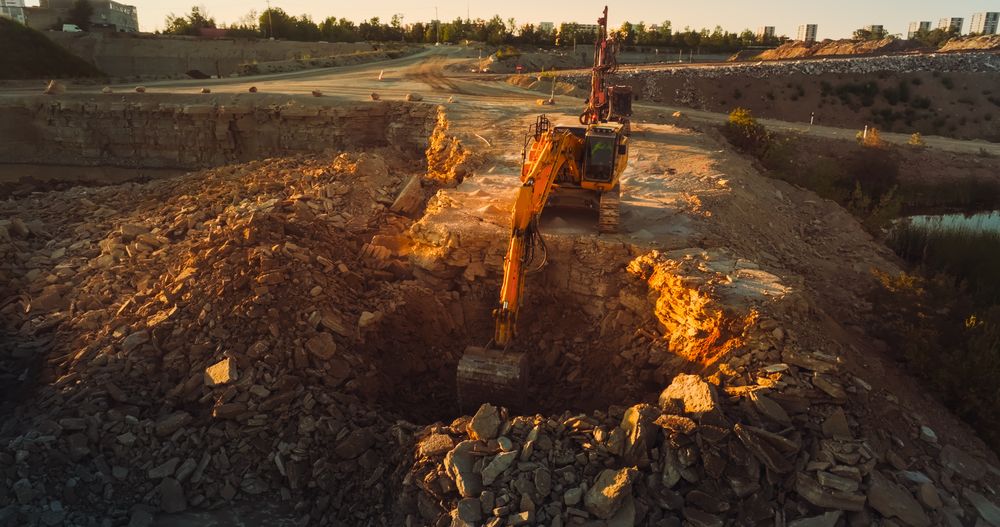 Aerial Drone Shot Of Construction Site On Sunny Day: Industrial Excavator Digging Rocks To Lay Foundation For Apartment Building. People Using Heavy Machinery To Complete A Real Estate Project.