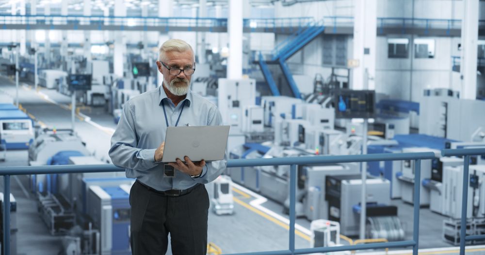 Factory Manager Working on Laptop at a Futuristic Industrial Manufacture with Autonomous AI Robots. Specialist Monitoring All the Data on a Computer, Checking Reports and Planning Workload