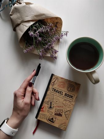 Cropped image of a hand holding a pen with "Travel book" and flower bouquet and cup of coffee on table