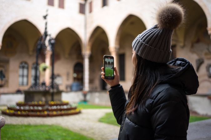 Content Asian female traveler in warm wear taking photo on smartphone while sightseeing at old city