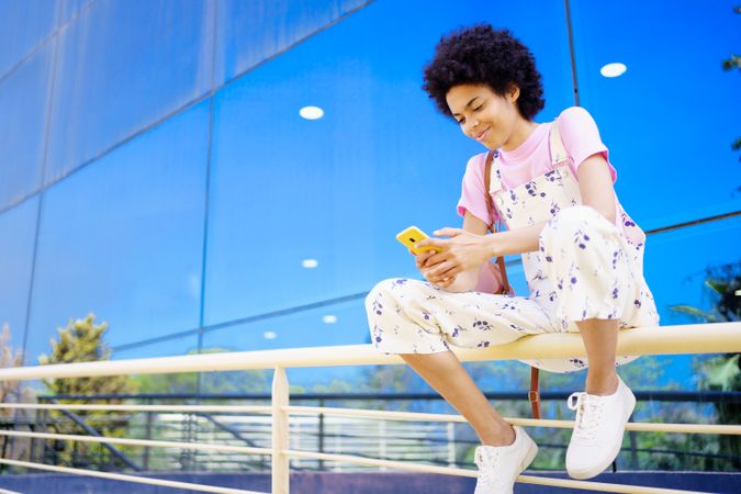 Smiling woman sitting on handrails in front of reflective building texting on phone