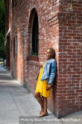 Full length shot of woman leaning against old brick building holding iced coffee looking at camera 5qkwKb
