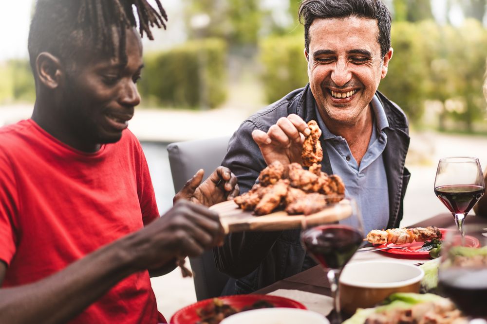 Happy multi-ethnic friends sitting at farmhouse table passing around meat dish