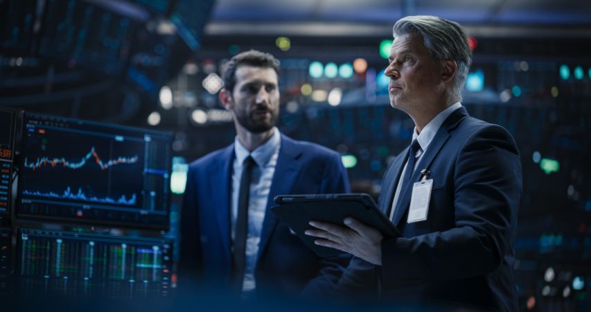 Team of Two Handsome Stock Exchange Brokers Using Tablet Computer, Chatting About the Asset Management Strategy for an International Business Client, Working in a Office Building with Computer Screens
