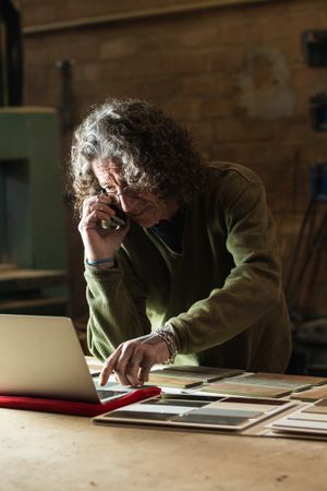Man talking on smartphone and typing on laptop in workshop