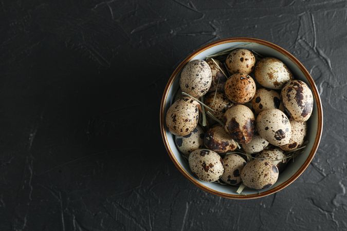 Many quail eggs in bowl on dark background, space for text and top view