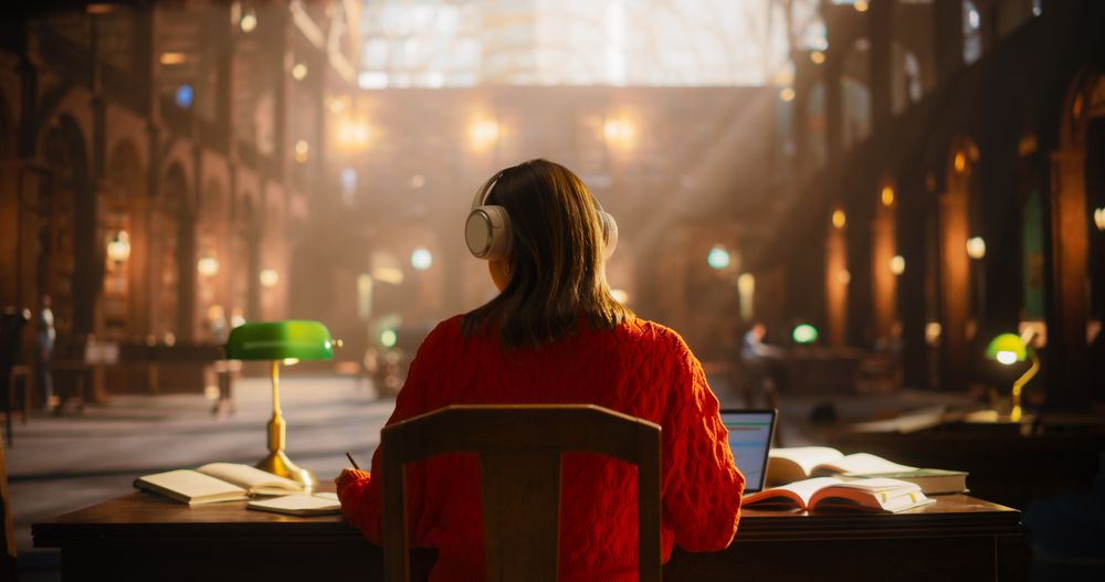 Young Woman in Red Sweater Engaged in Studying at a Classical Library, Using Headphones and Laptop for E-Learning