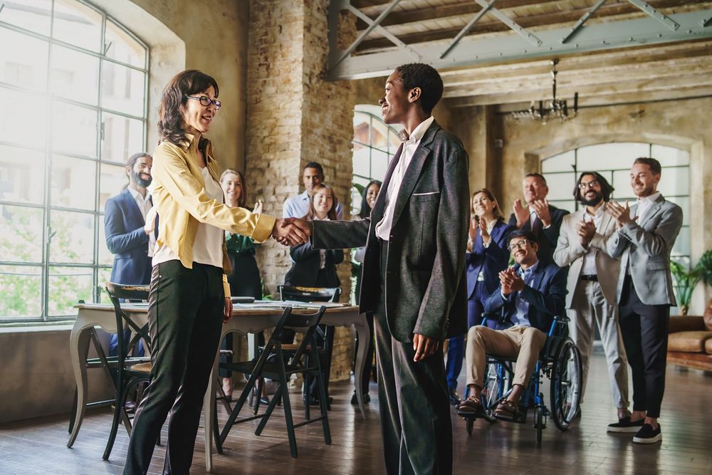 Multi-ethnic businesswomen shaking hands in front of their colleagues applauding in background