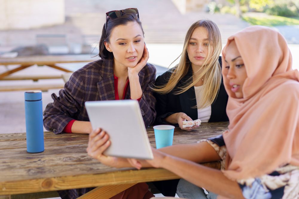Three friends on park bench with insulated mugs looking at something on digital tablet