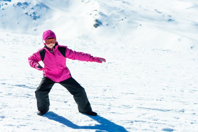 Child in pink snow suit practicing snow board poses on snowy mountain