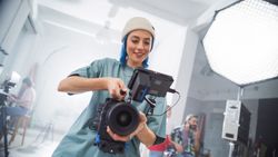 Young Female Filmmaker In A Beanie Adjusts A Camera On A Busy Film Set, Capturing The Essence Of Modern Cinematography With Her Team In The Background, Focused On A Creative Project.